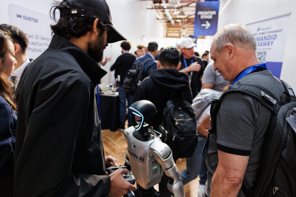 Attendee using a handheld controller to operate a small humanoid robot powered by PopUp WiFi on the crowded AUTONOMOUS 2026 expo floor, with the Frontier Stage sign visible.