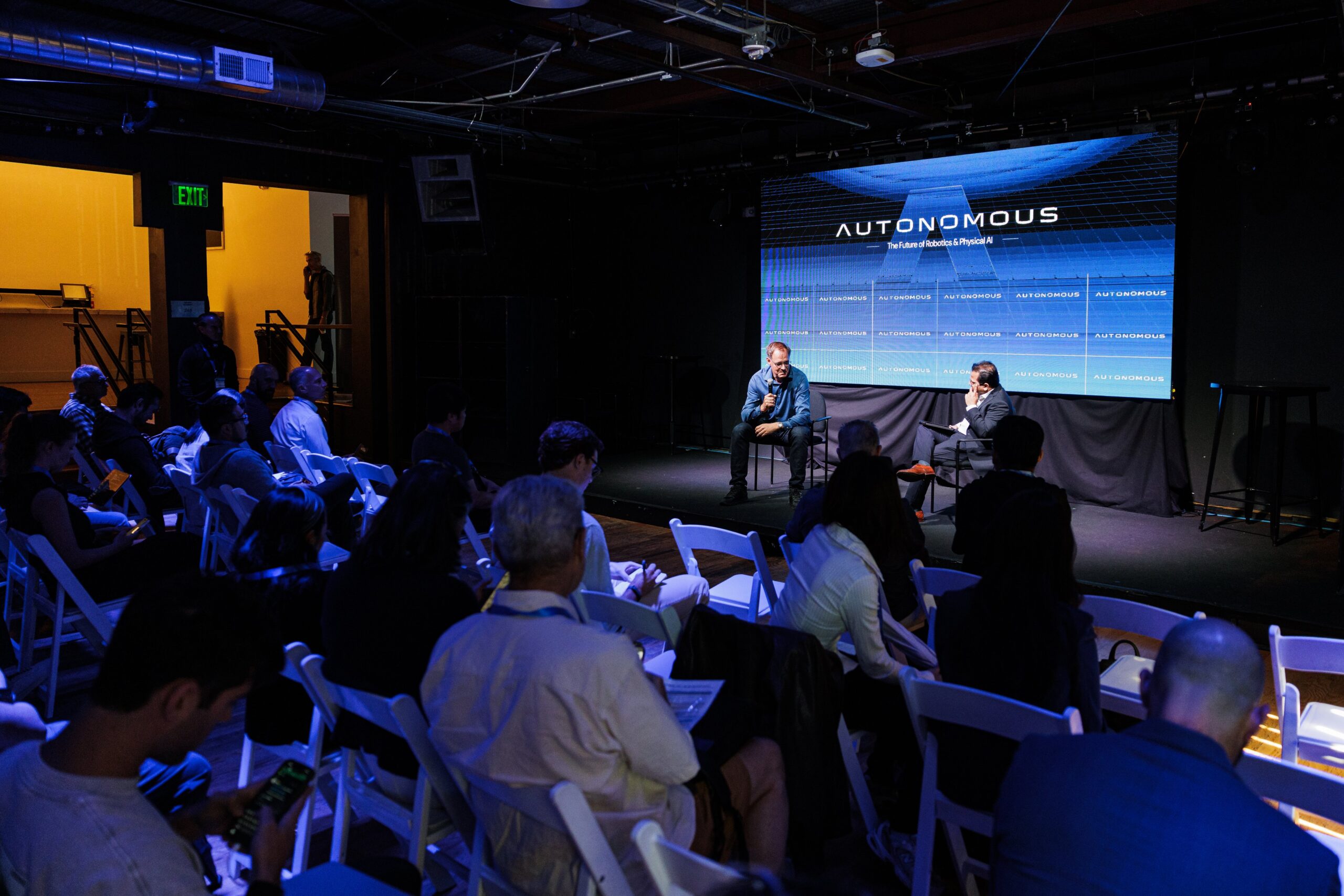 Speakers seated on stage for a fireside chat in front of the AUTONOMOUS "Future of Robotics and Physical AI" screen, with a seated audience at The Midway in San Francisco