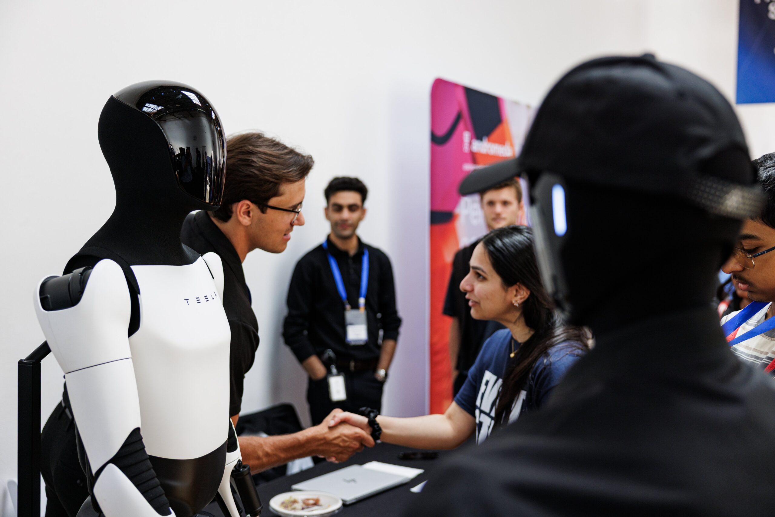 Two attendees shaking hands beside a Tesla Optimus humanoid robot at the AUTONOMOUS 2026 robotics and physical AI conference