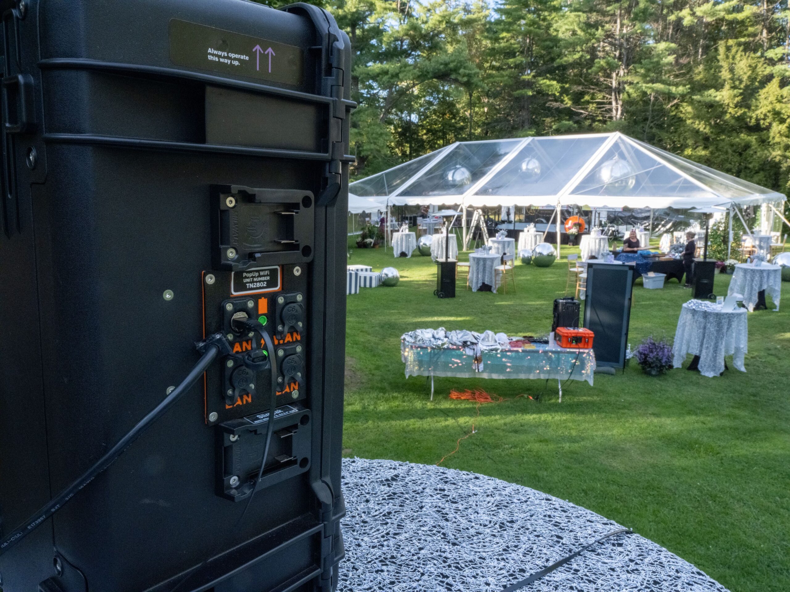 A PopUp WiFi unit's network ports in close-up, with a clear-top tent and cocktail tables in a pine-lined field at Storrs Pond, New Hampshire