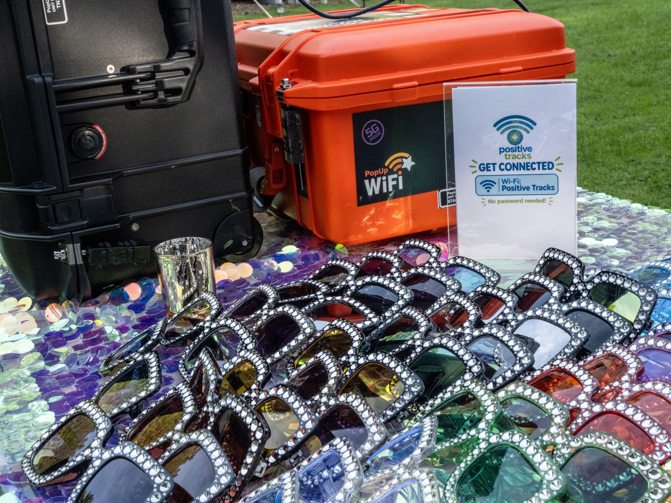 PopUp WiFi event WiFi rental units on a sequin table beside a Positive Tracks WiFi sign and rows of rhinestone sunglasses