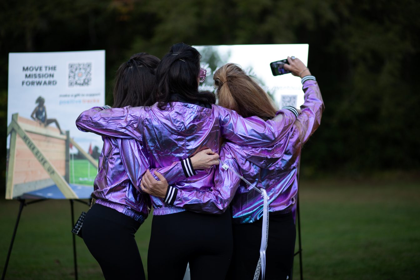 Three guests in matching purple metallic jackets taking a selfie beside a Positive Tracks QR code donation sign in New Hampshire