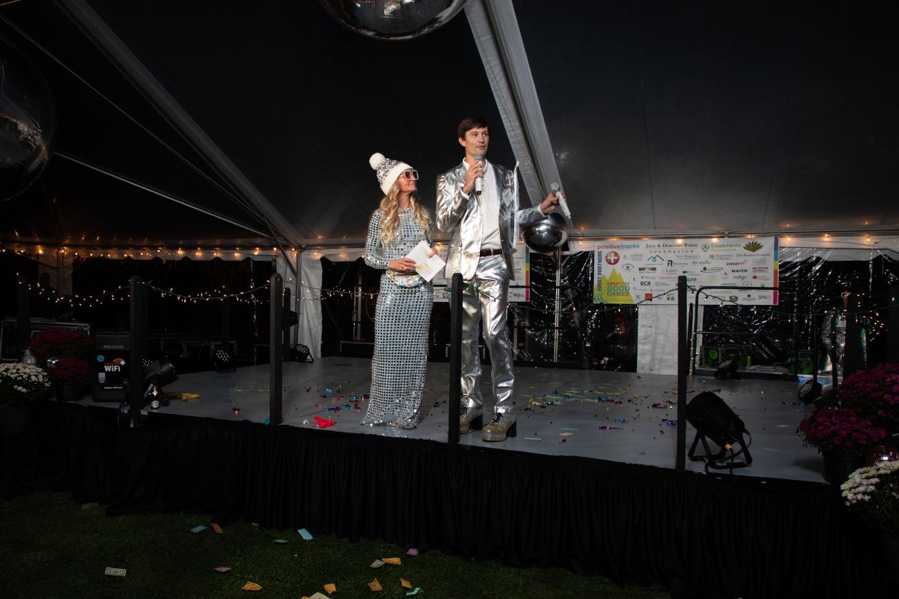 Two hosts in silver outfits on a tented stage at Sweat For Good Games in Hanover, New Hampshire, with a PopUp WiFi unit side stage